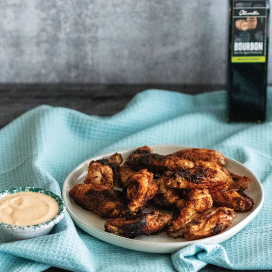 Grilled chicken wings on a white plate with a side of creamy dipping sauce, set on a light blue textured cloth with a bottle of Bourbon sauce in the background.