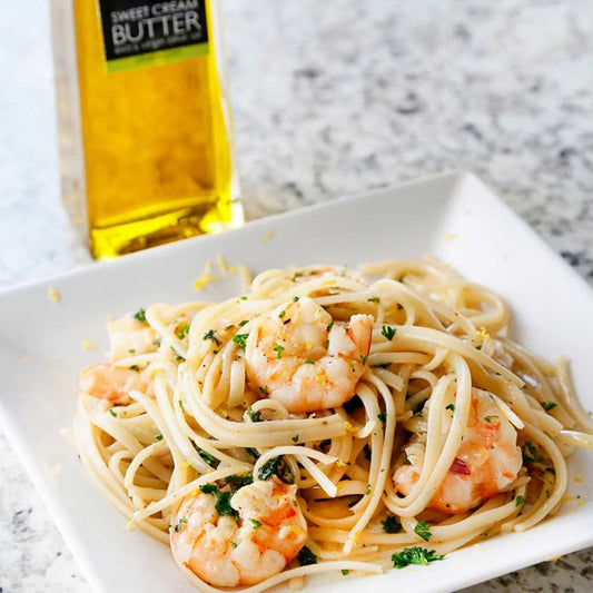 A close-up of a plate of spaghetti with shrimp and herbs, with a bottle of sweet cream butter in the background on a speckled countertop.