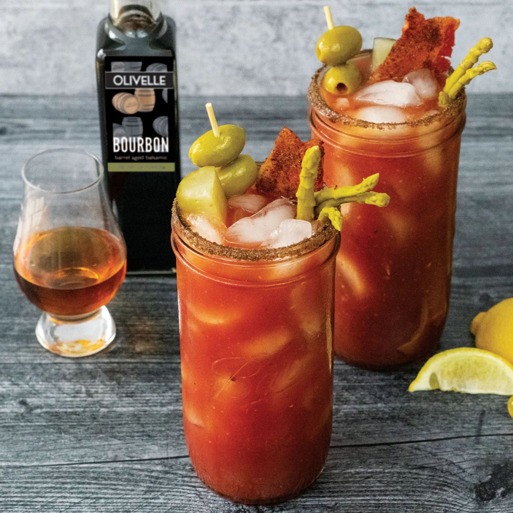 Two Bloody Mary cocktails with olives and chips, a glass of bourbon, and lemons on a wooden surface.